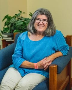 Image of Rosemary Schwoerer- a white woman with shoulder length hair and a blue shirt. She is staging in a chair and smiling. The background is beige with a small plant.